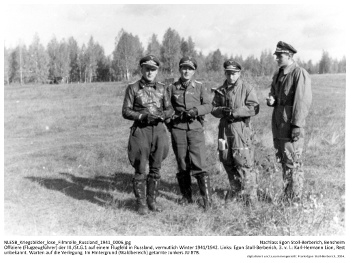 Offiziere (Flugzeugführer) der III./St.G.1 auf einem Flugfeld in Russland, vermutlich Winter 1941/1942. Links: Egon Stoll-Berberich, 3. v. l.: Karl-Hermann Lion, Rest unbekannt. Warten auf die Verlegung. Im Hintergrund (Waldbereich) getarnte Junkers JU 87B. NLESB_Kriegsbilder_lose_Filmrolle_Russland_1941_0006