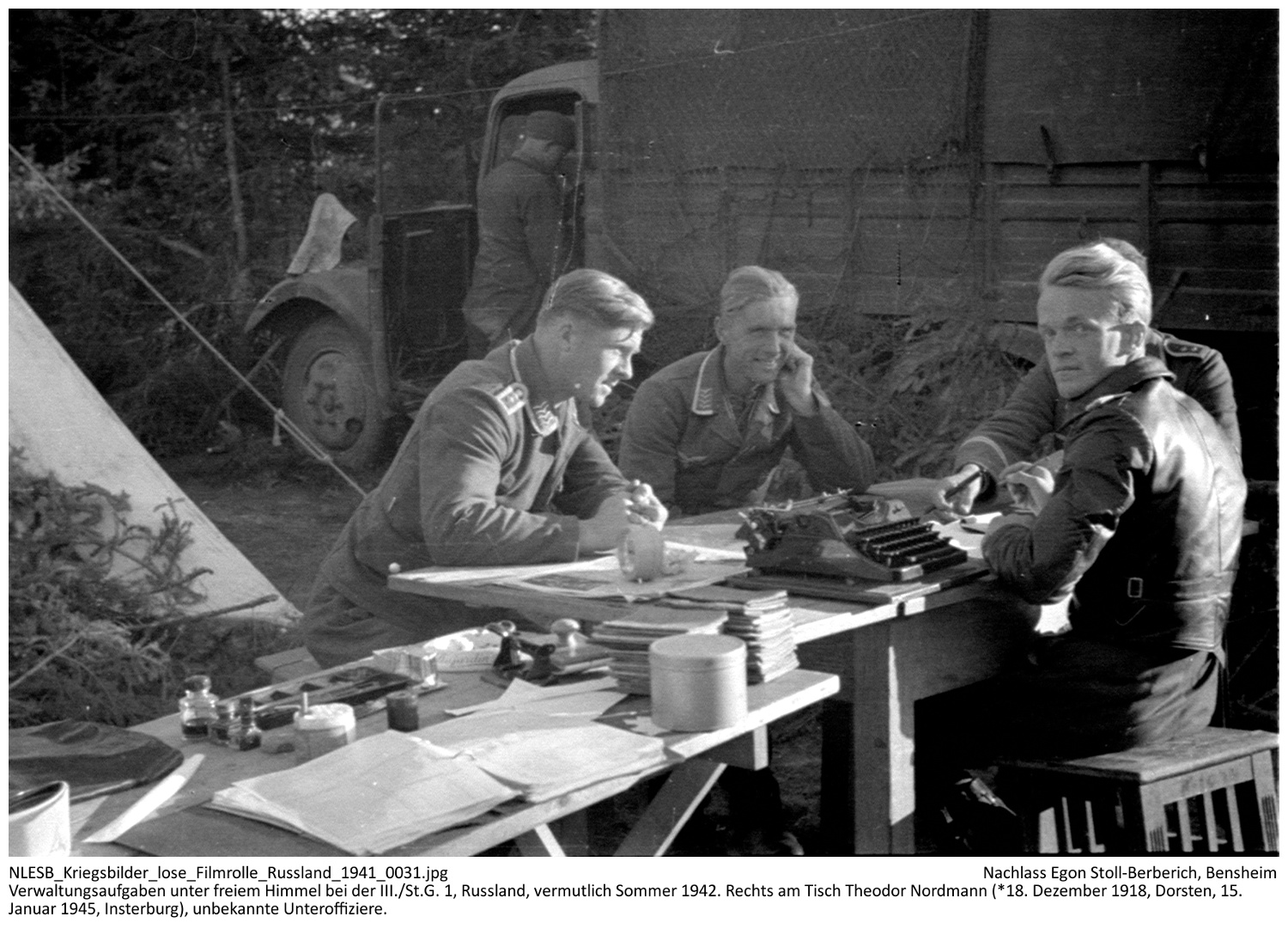 Verwaltungsaufgaben - hier Abzeichnen der Leistungsbücher (Stapel auf dem vorderen Tisch) - unter freiem Himmel bei der III./St.G. 1, Russland, vermutlich Sommer 1942. Rechts am Tisch Theodor Nordmann (*18. Dezember 1918, Dorsten, 15. Januar 1945, Insterburg), unbekannte Unteroffiziere. NLESB_Kriegsbilder_lose_Filmrolle_Russland_1941_0031