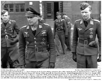 Hauptmann Egon Stoll-Berberich (2. v. l.) beim Antreten der III./St.G.1 in Wilna, vermutlich April 1944. Rechte Brusttasche: Deutsches Kreuz in Gold (17. April 1943), Am Hals: Ritterkreuz des Eisernen Kreuzes (29. Februar 1944), oberhalb der linken Brusttasche: Schlachtfliegerabzeichen mit Stern (12. August 1943), darunter Bandspange mit Eisernem Kreuz 2. Klasse (1. November 1939), Dienstauszeichnung IV. Klasse (15. April 1938), Medaille zur Erinnerung an den 13. März 1938, Medaille zur Erinnerung an den 1. Oktober 1938 mit Spange "Prager Burg" (1. Oktober 1939). Auf der linken Brusttasche: Eisernes Kreuz 1. Klasse (11. Juli 1940). Unter der linken Brusttasche: Flugzeugführerabzeichen (6. Dezember 1938) und Verwundetenabzeichen in Schwarz (8. April 1943). NLESB_Kriegsbilder_lose_Wilna_0007